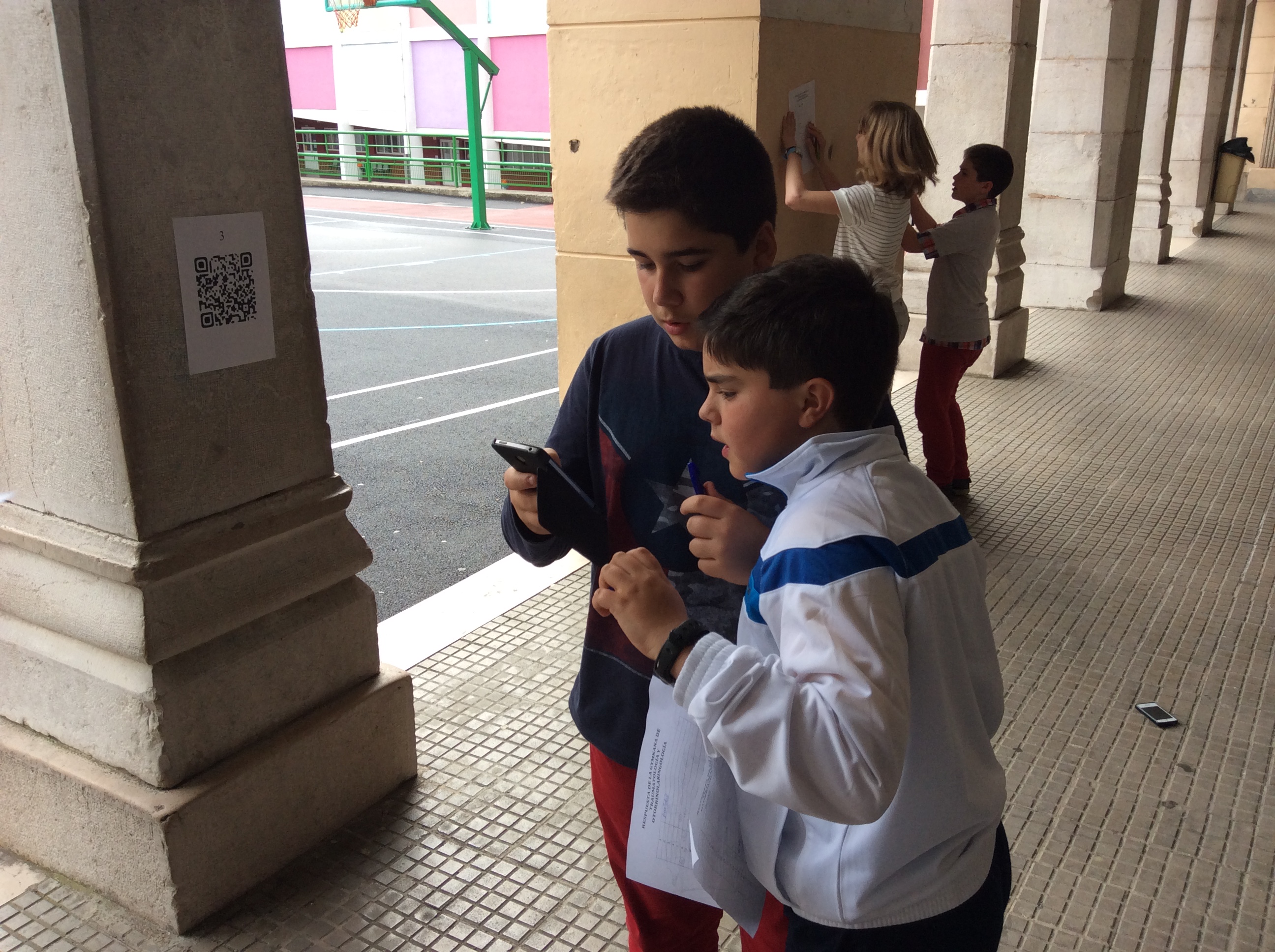 Dos adolescentes usando un smartphone bajo una arcada, con un código QR visible en un pilar y una cancha de baloncesto detrás.